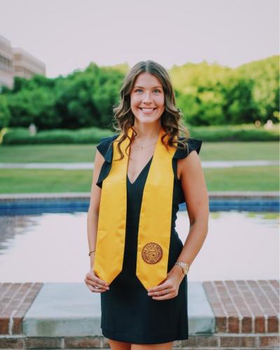 A woman in a black dress and yellow graduation sash stands in front of a brick wall and a pool.
