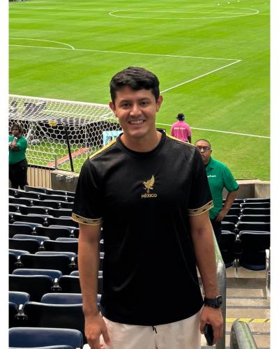 A man smiles while standing in bleachers overlooking a soccer field.