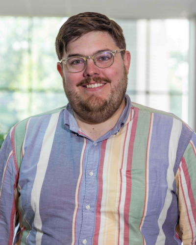 A man with short brown hair and glasses smiles while wearing a colorful, striped button-up shirt.