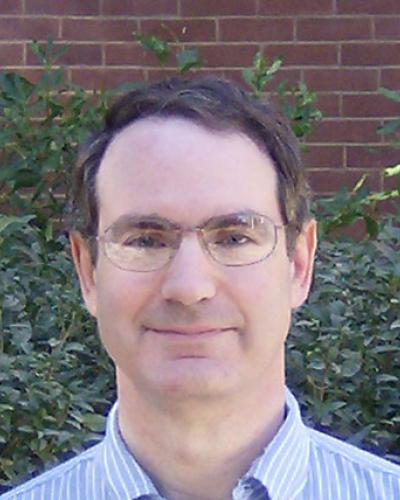 A man wearing glasses and a blue and white striped shirt smiles at the camera in front of a brick wall and some greenery.