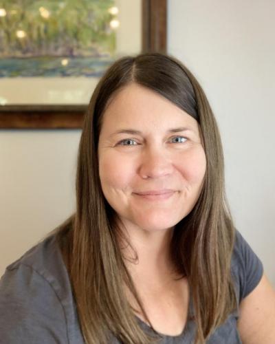 A woman with long brown hair and a gray t-shirt smiles at the camera in front of a painting on a white wall.