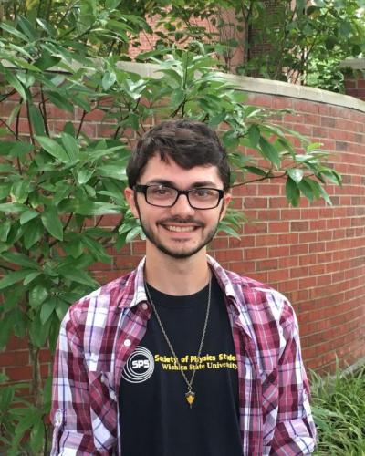 A man stands in front of a brick wall and greenery, smiling and wearing a plaid shirt, black t-shirt, glasses, and a necklace.