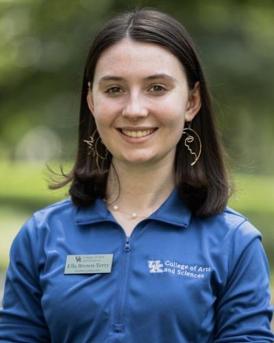 A woman smiles while wearing a blue shirt with the University of Kentucky College of Arts and Sciences logo.