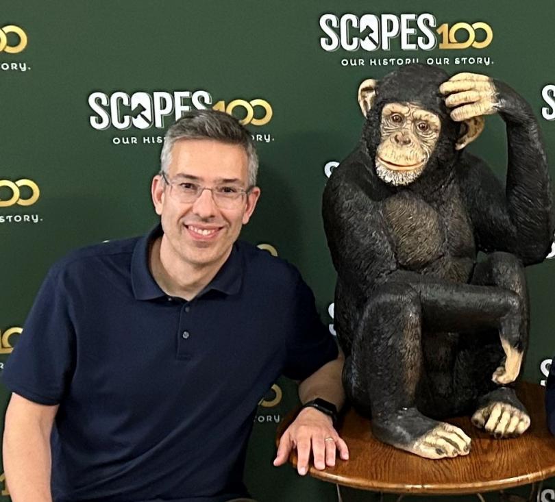Dr. Antonis Rokas smiling with a Scopes 100 backdrop in the background with a large chimpanzee figurine sitting next to him on a wooden desk stand.